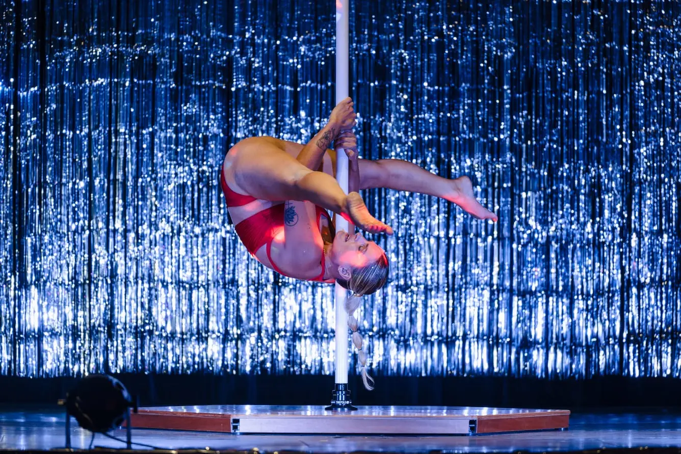 Danseuse suspendue au pôle, jambes repliées, devant un rideau bleu scintillant