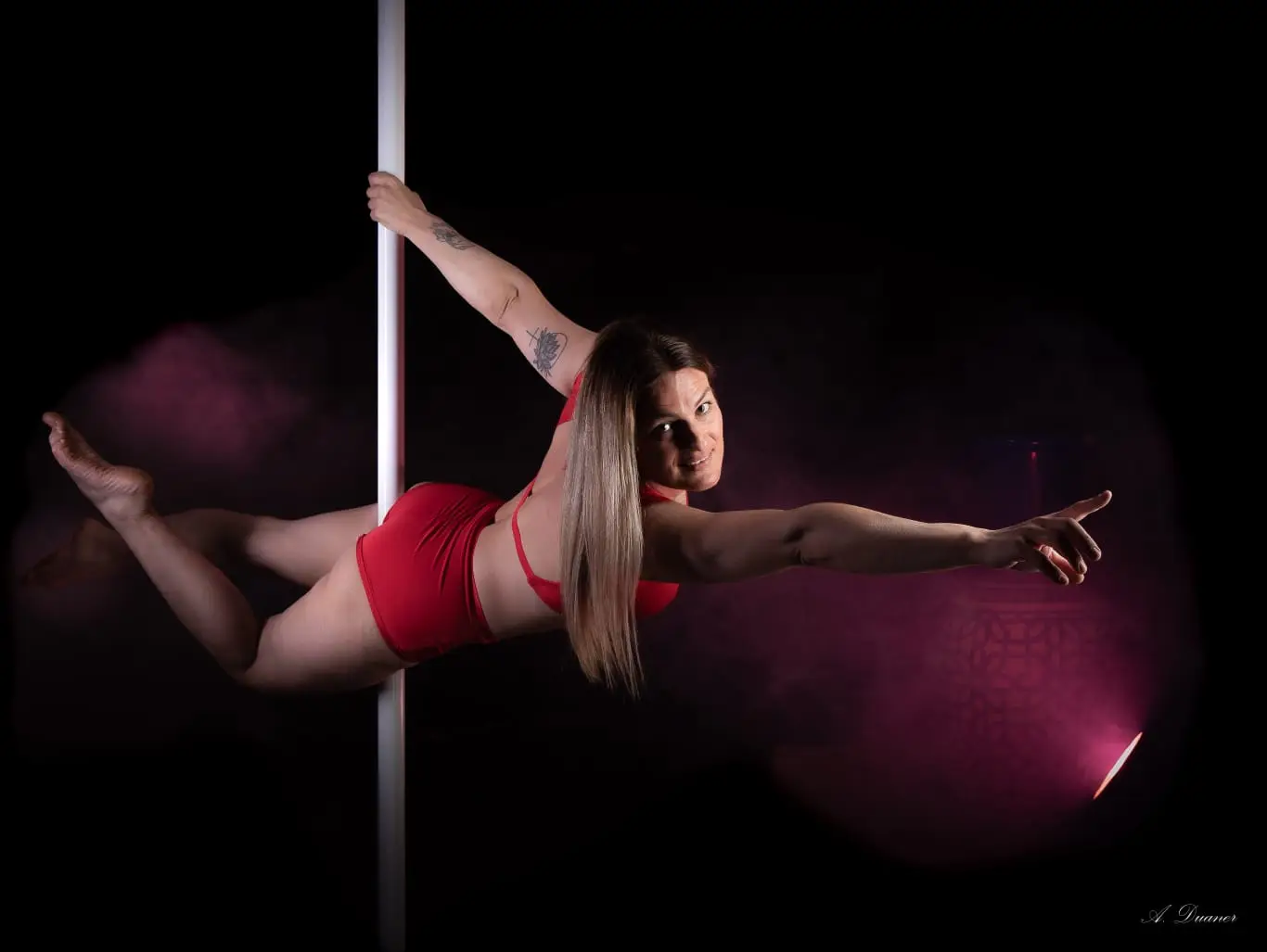 Une danseuse en tenue rouge, à l’horizontale au pôle, dans une brume rose éclairée