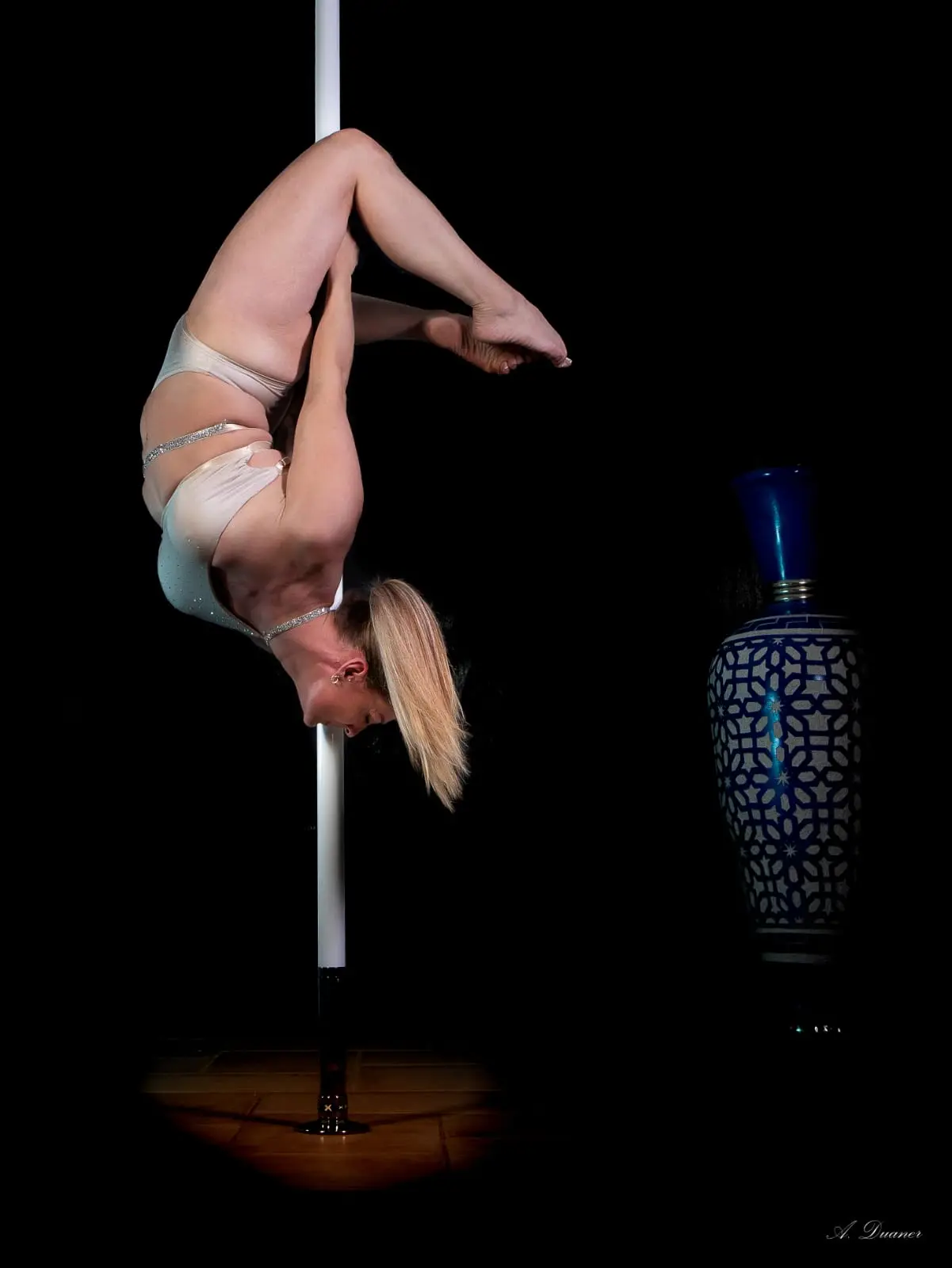 Danseuse en figure inversée le long du pôle, corps replié, en tenue champagne, sur fond noir